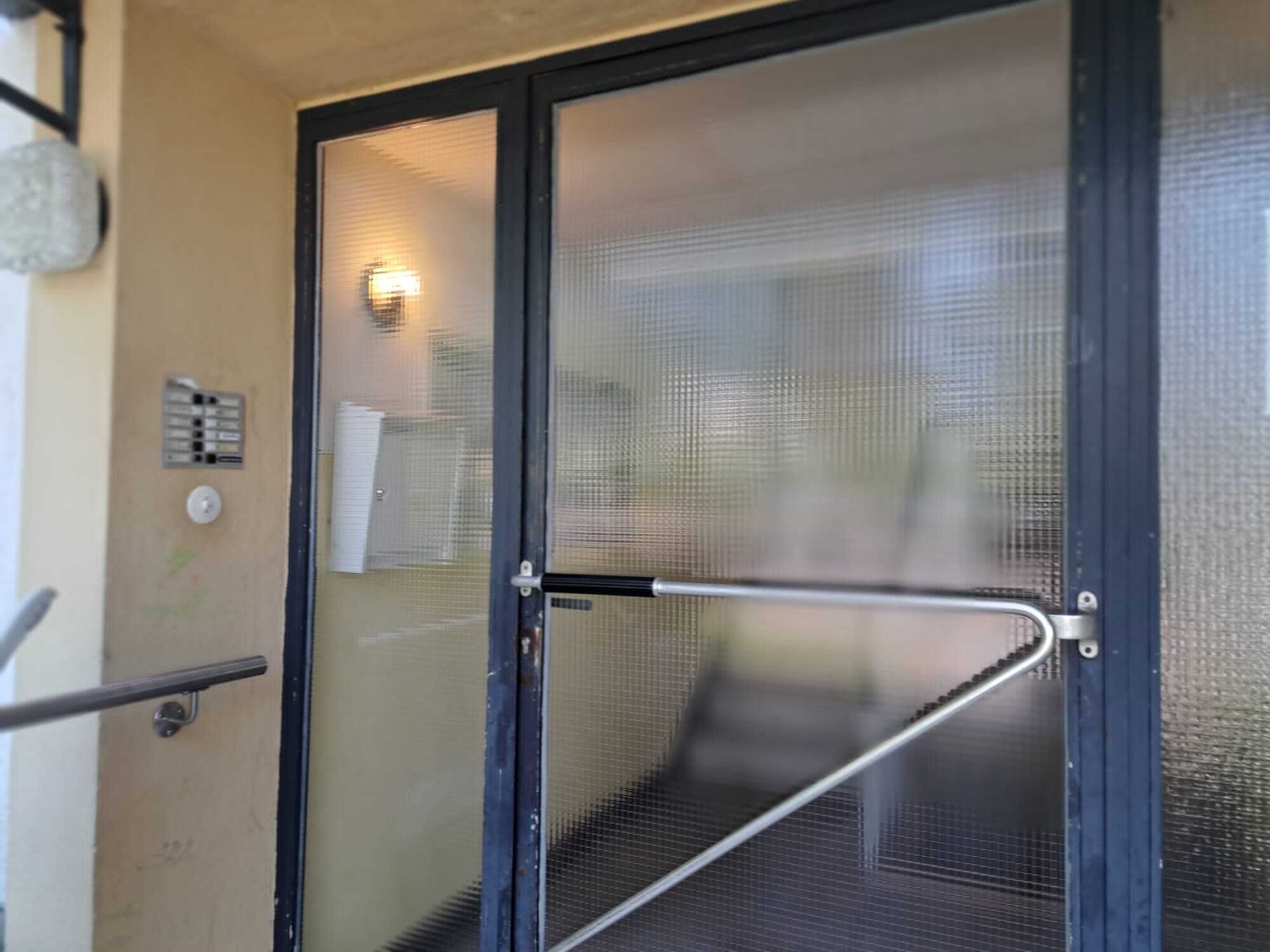 Una puerta de vidrio con una barra de seguridad metálica, un portero automático en la pared y un pequeño ventanal.