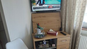 A room with a television displaying a news program, a small wooden cabinet with drawers, a white chair, and a radiator. - 5 km from the center, 1 km from the train station and 11 km from Baden-Airpark (Baden-Baden)