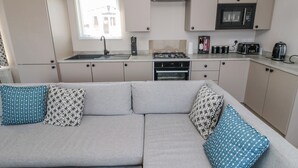 A compact kitchenette with a gas stove, oven, and microwave. A grey sofa with patterned cushions is in the foreground.
