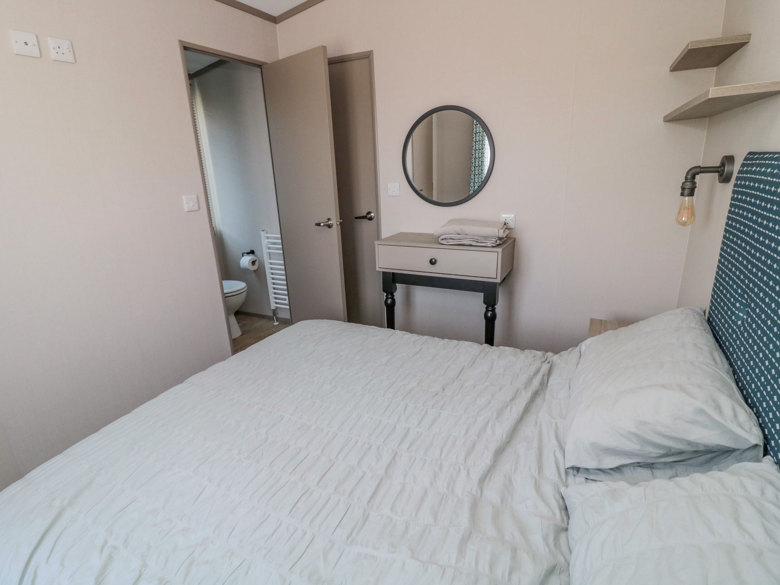 A bedroom with a bed, a bedside table with a mirror, a radiator, and a door leading to a bathroom.