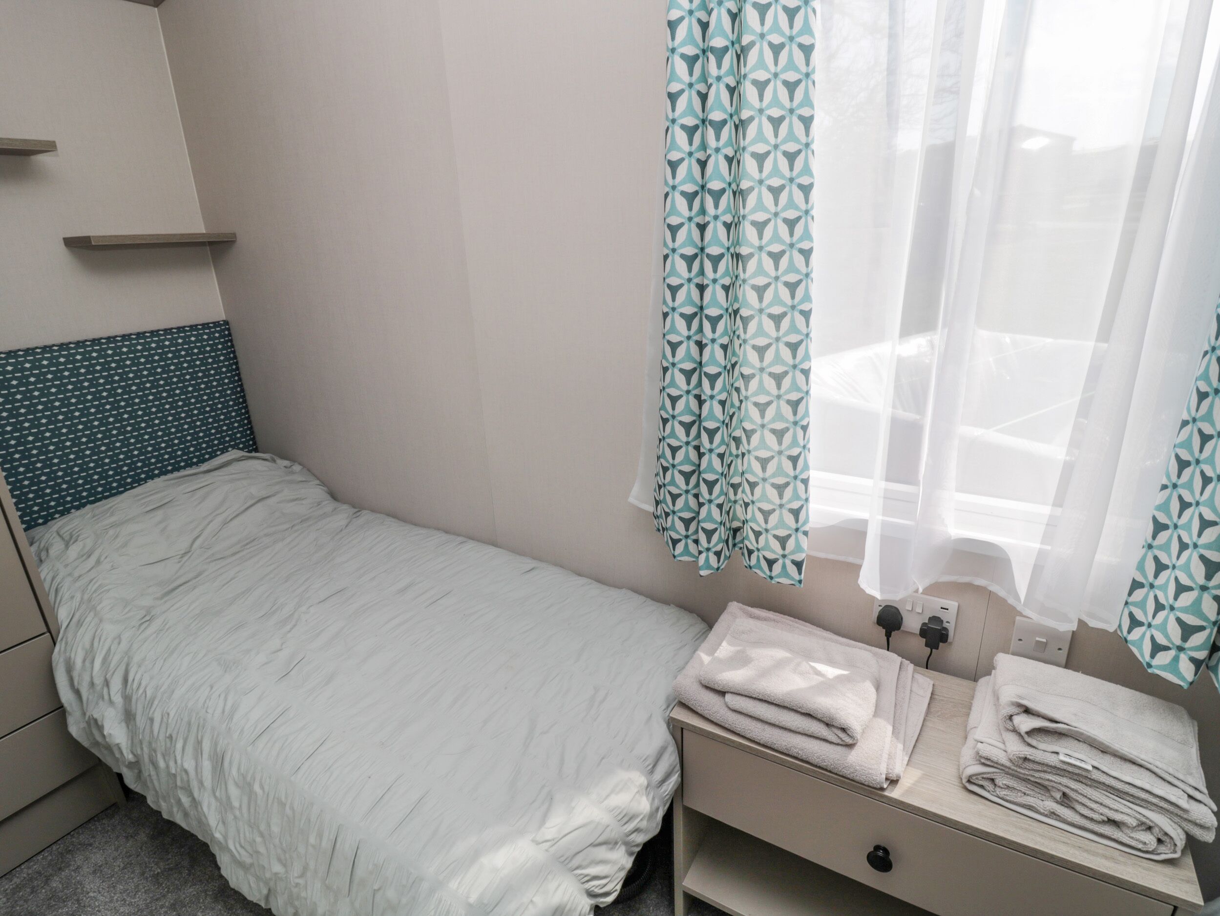 A single bed with a patterned headboard, a bedside table with towels, and a window with patterned curtains.