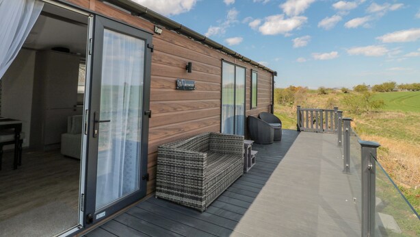 A modern wooden cabin with a glass door entrance, a deck with outdoor seating, and a sign indicating the name 'Beckwood'.