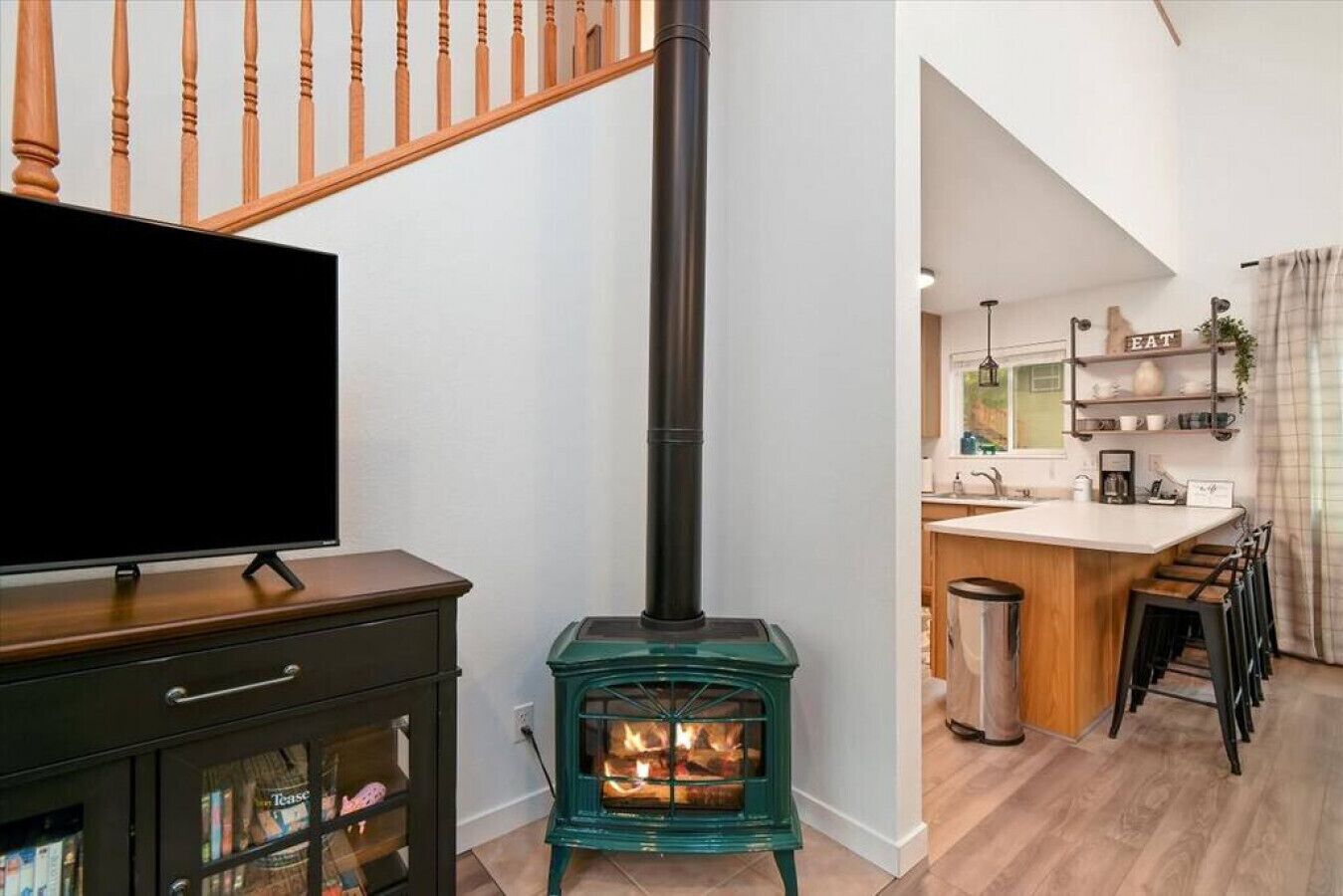 A modern living room with a television, a green wood-burning stove, a wooden staircase, and a kitchen area with a bar stool and a trash can.