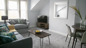 En moderne stue med en grå sofa, et sofabord med en skål frugt, et fjernsyn på et træstel og et spiseområde med en stol og en urtepotte med planter.