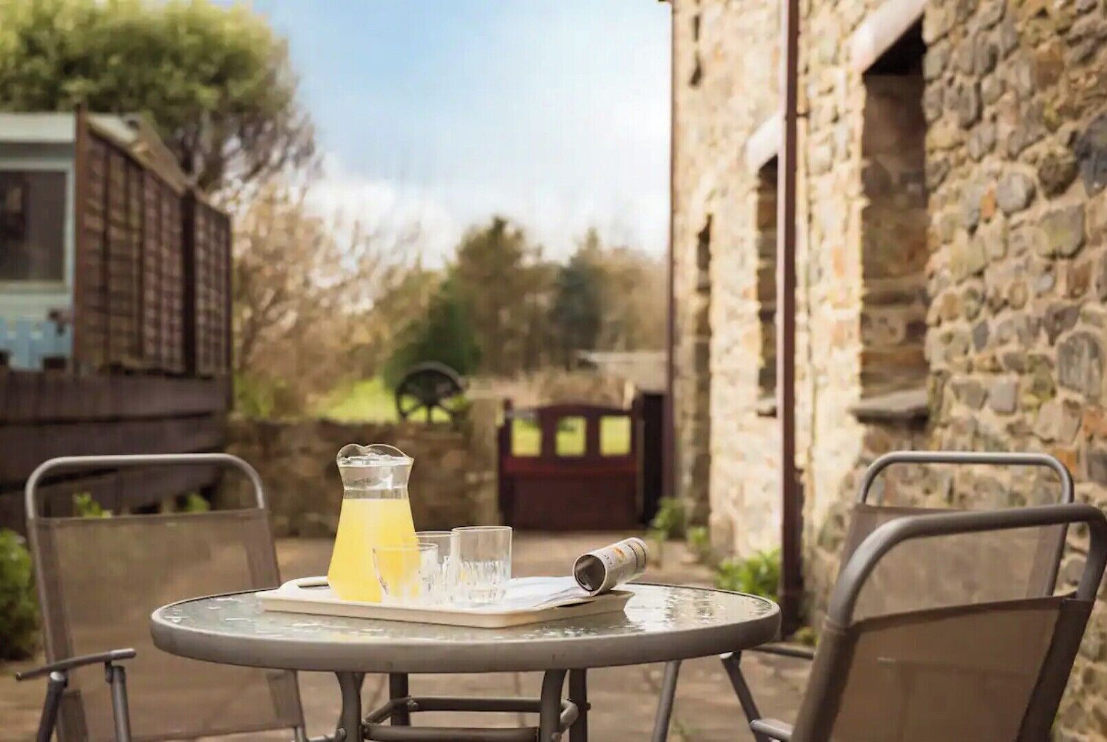 A table with a pitcher of yellow liquid, two glasses, and a rolled-up menu in an outdoor setting with stone walls and a wooden gate in the background.