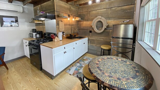 Een kleine keuken met een centraal kookeiland, een kookplaat, een magnetron en een koelkast. Er staat een ronde tafel met krukken en aan de muur hangt een decoratieve spiegel.
