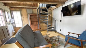 Een woonkamer met een houten plafond, een trap, een aan de muur bevestigde televisie en een blauwe fauteuil.