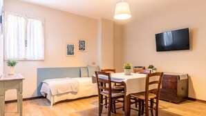 A cozy room with a bed, a dining table with chairs, a TV mounted on the wall, and a window with curtains.