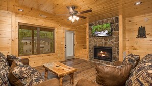 A cozy log cabin interior with a stone fireplace, a ceiling fan, and a television mounted on the wall.