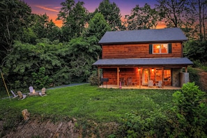 A two-story wooden cabin with a porch, surrounded by lush greenery. - Secluded 3 King Suite Gatlinburg Cabin | Swim Spa+Hot Tub, Games & Flat Parking (Gatlinburg)