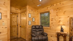 A wooden cabin interior with a leather armchair, a small table with a lamp, and framed pictures on the wall.