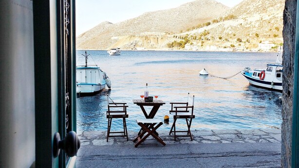 A view from a window overlooking a harbor with boats, a table with chairs, and a mountainous backdrop. - SeaMe House I (Symi)