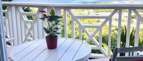 A round white table with a potted plant on a wooden deck with a white railing, overlooking a coastal view.