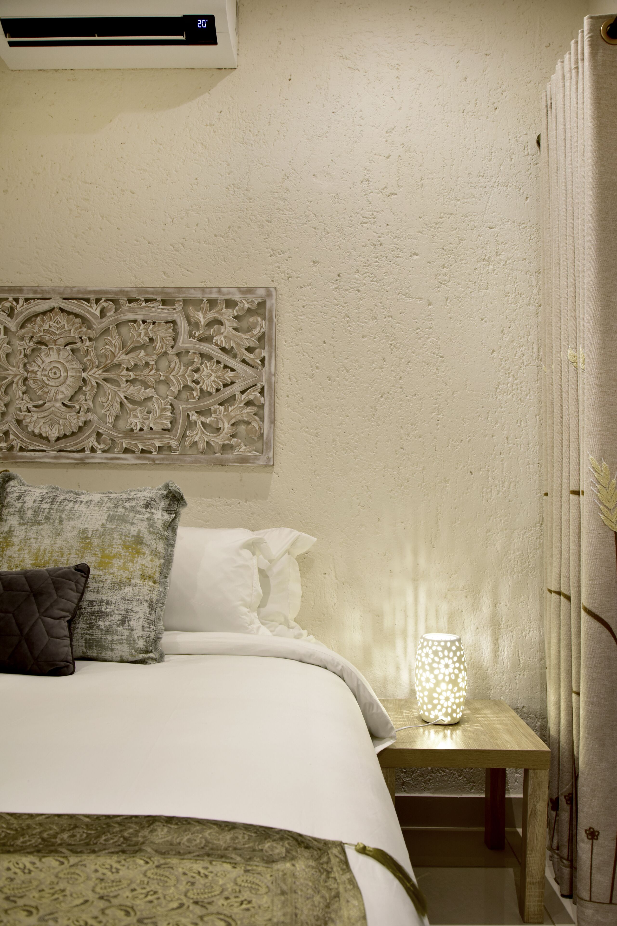 A neatly made bed with a decorative headboard, a bedside table with a lamp, and a wall-mounted air conditioner.