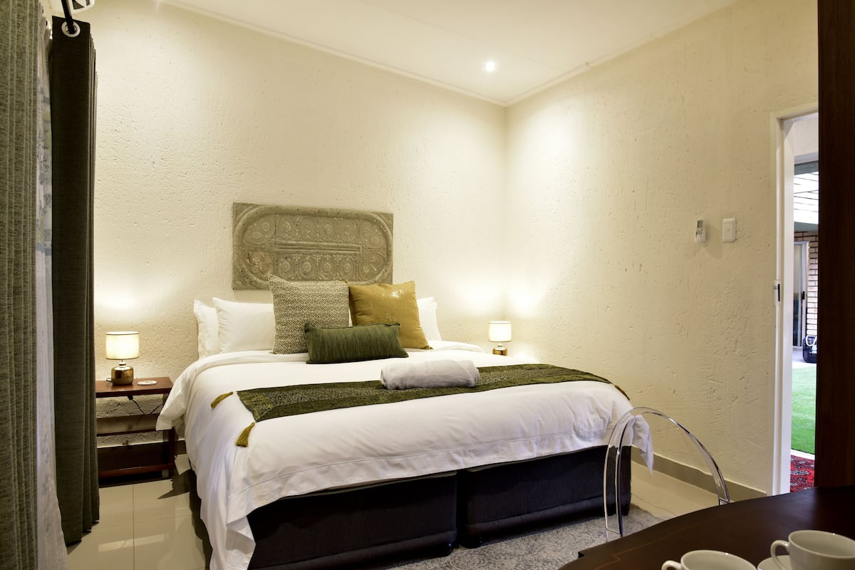 A neatly arranged hotel room with a large bed, two bedside tables with lamps, a headboard with a decorative panel, and a view of the outdoors through a door.