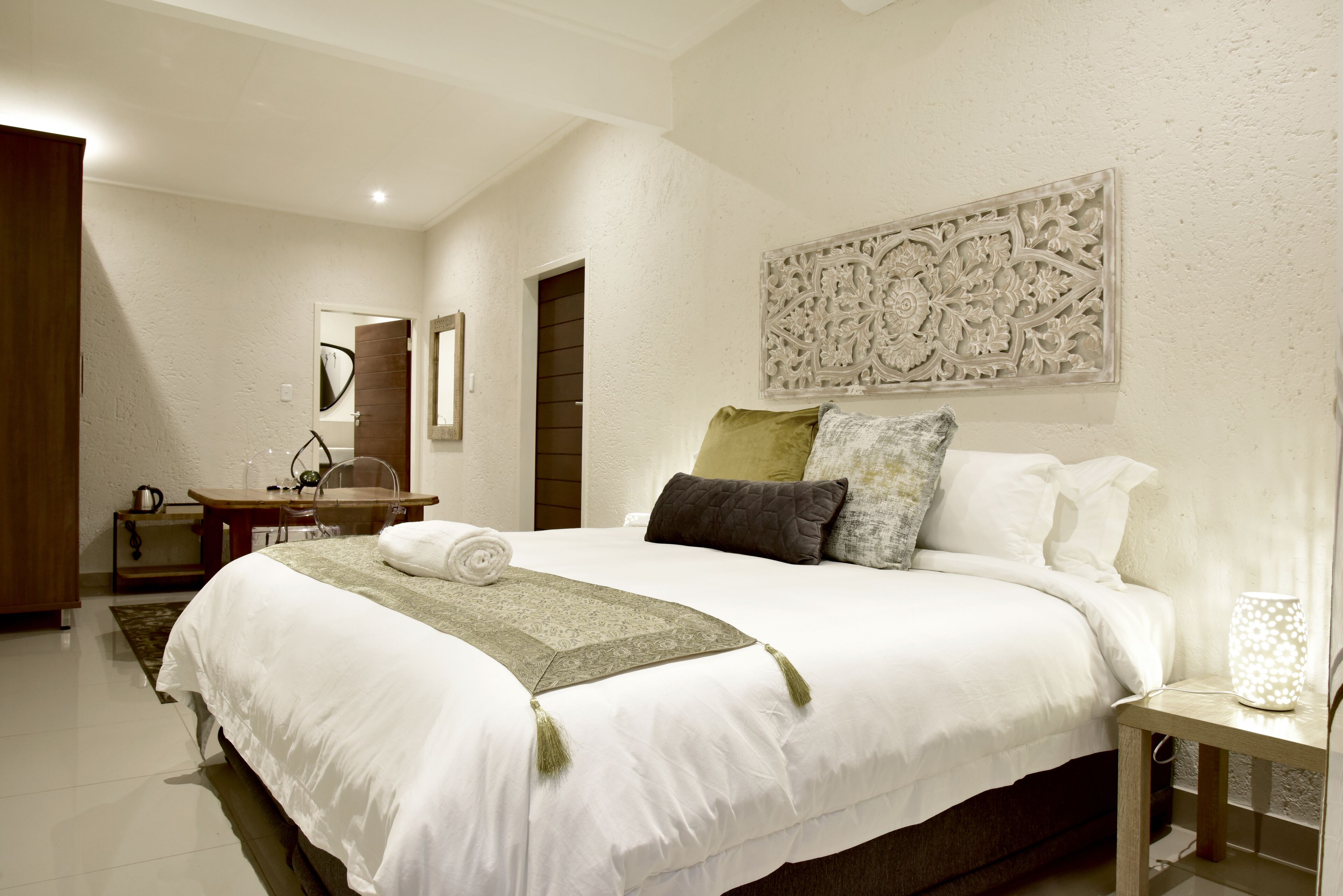 A neatly arranged bedroom with a large bed, a bedside table, a mirror, and a decorative wall piece.