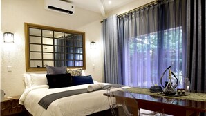 Une chambre d’hôtel avec un grand lit, une tête de lit en bois, un miroir, une fenêtre avec des rideaux, un bureau avec une chaise et un vase décoratif.