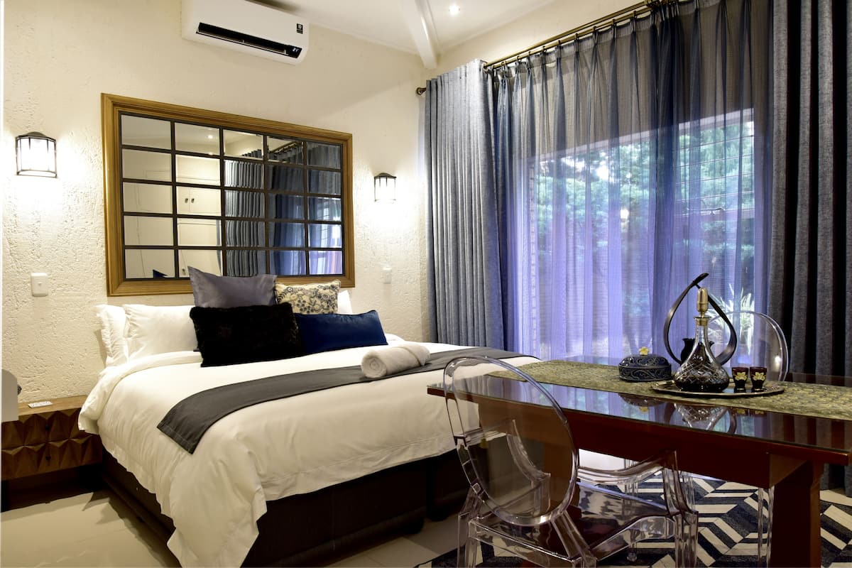 A hotel room with a large bed, a wooden headboard, a mirror, a window with curtains, a desk with a chair, and a decorative vase.