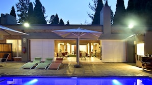 Un espace piscine avec des chaises longues et un parasol, une terrasse couverte avec des tables et des chaises, et un bâtiment en briques doté d’une porte de garage.