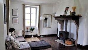 Ein gemütliches Wohnzimmer mit Kamin, einer Couch, einem kleinen Tisch mit einer Vase und einem Fenster mit Vorhängen.