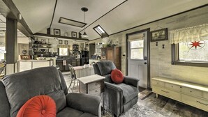 A spacious living room with a grey sofa, a matching armchair, and a small table with drawers. A window with a lace curtain and a decorative starburst window treatment is visible.