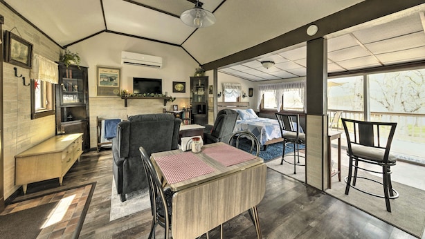 A spacious living area with a dining table, chairs, a sofa, and a fireplace. There is a kitchen area with a refrigerator, stove, and cabinets. The room has large windows and a ceiling fan.