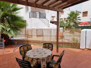 Een terras buiten met een ronde tafel en stoelen, een bamboe hek en een wit gebouw op de achtergrond.