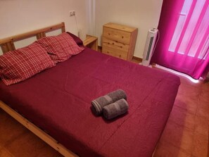 Een bed met een rode deken, twee kussens en twee opgerolde handdoeken. Er staat een houten nachtkastje met een lade, een klein tafeltje en een witte airconditioner.