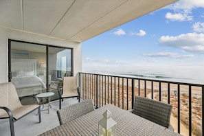 A balcony with a view of the ocean, furnished with a table and chairs, and a glass door leading to a bedroom.