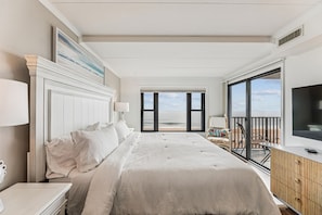 A bedroom with a large bed, a TV, a balcony with a view of the beach, and a painting on the wall.