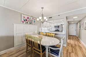 Zona pranzo con tavolo e sedie bianche, un lampadario e una cucina sullo sfondo.