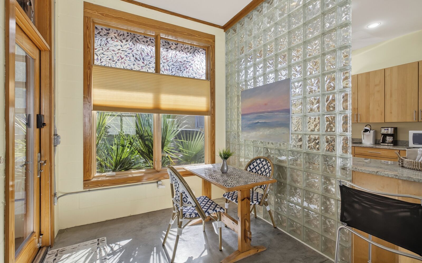 A compact kitchen with a dining area, featuring a window with blinds, a decorative glass partition, and wooden cabinets.