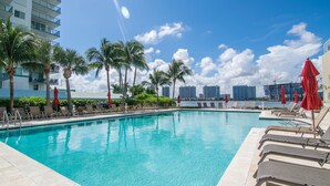 Una piscina con sillas de descanso y sombrillas, rodeada de palmeras y un horizonte urbano al fondo.