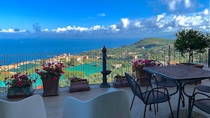 Terraço com mesa e cadeiras, plantas em vasos e vista para o mar e paisagem.