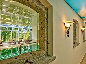 A hotel corridor with a glass-enclosed pool area, stone walls, and wall-mounted lighting fixtures. - Aquaquell Bad Pilzweg Hotel & Spa (Fuerstenzell)