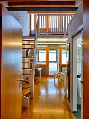 A spacious loft-style room with a wooden staircase leading to an upper level, a dining area with a table and chairs, and a view through large windows. - Aquaquell Bad Pilzweg Hotel & Spa (Fuerstenzell)