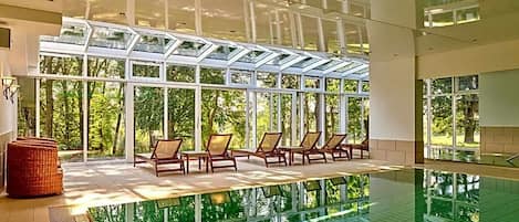 Una piscina interior con techo de cristal, sillones de descanso y vistas a zonas verdes.