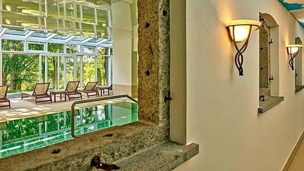 A hotel corridor with a glass-enclosed pool area, stone walls, and wall-mounted lighting fixtures. - Aquaquell Bad Pilzweg Hotel & Spa (Fuerstenzell)