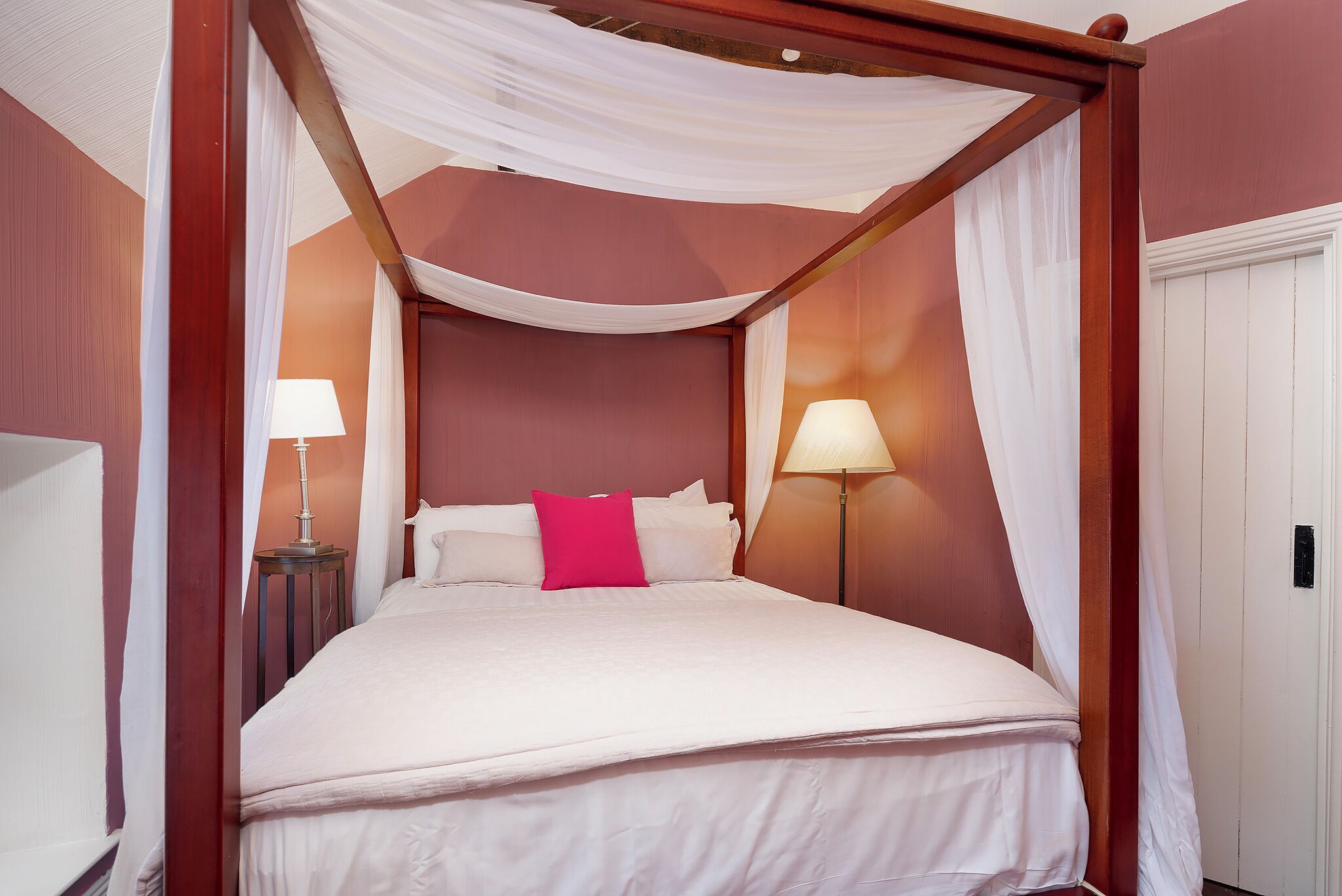 Una cama con dosel tapizada con sábanas blancas y una única almohada rosa brillante, con dos lámparas de noche a cada lado.