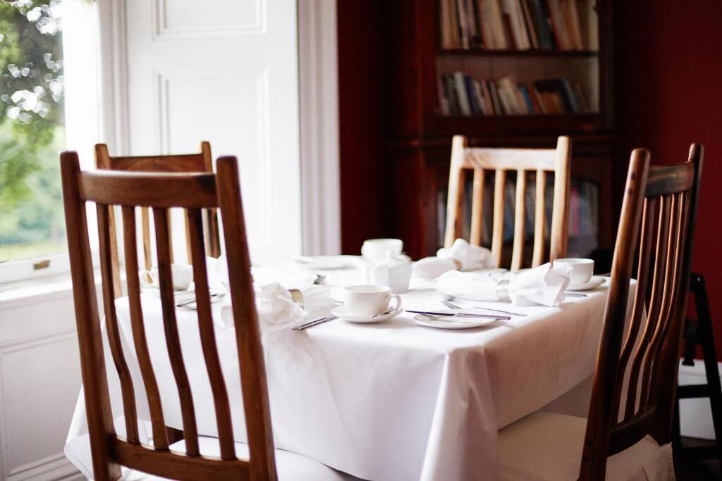 A dining table set with white tablecloth, wooden chairs, and a bookshelf in the background.