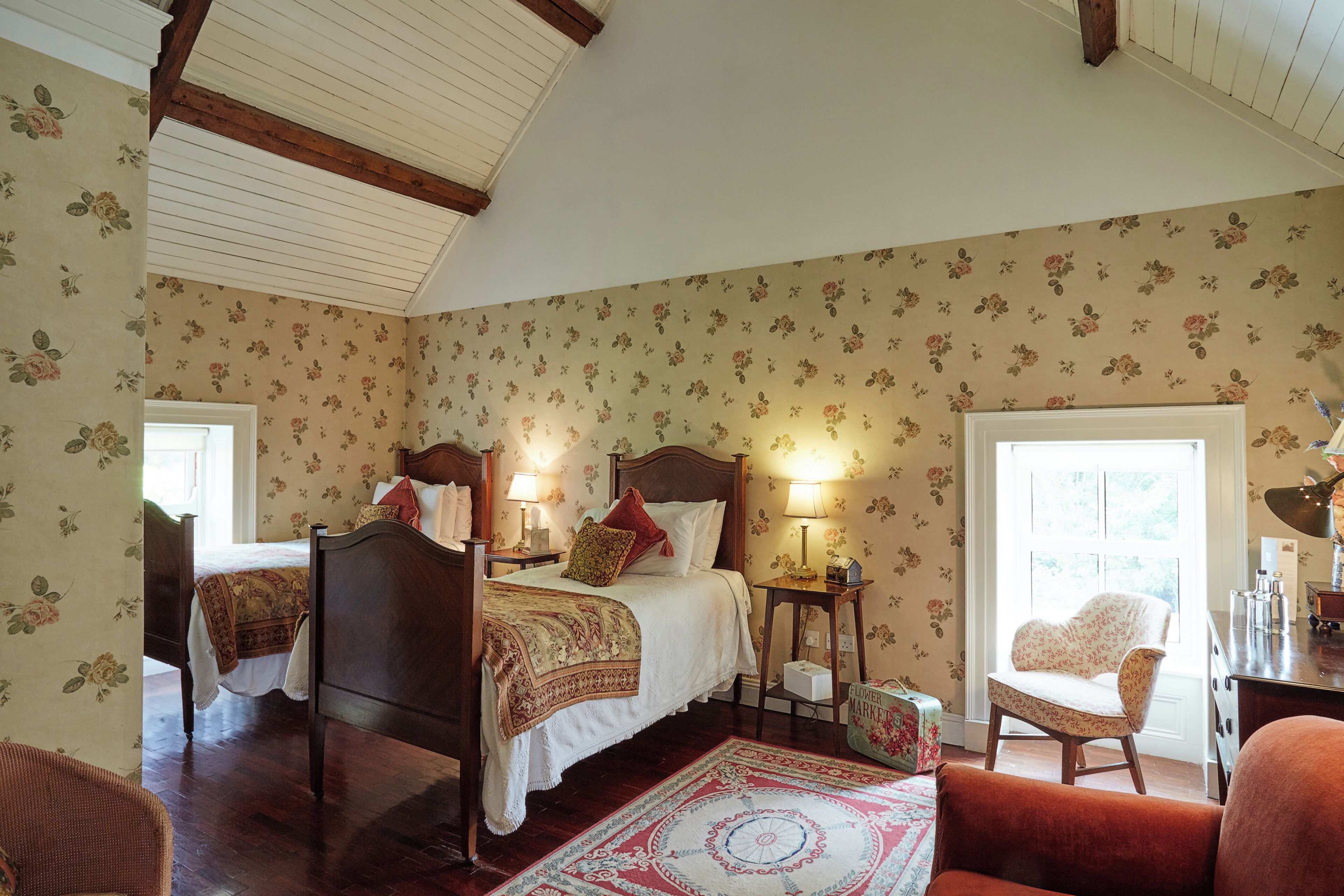 A bedroom with two beds, a wooden ceiling, floral wallpaper, and a red patterned rug.