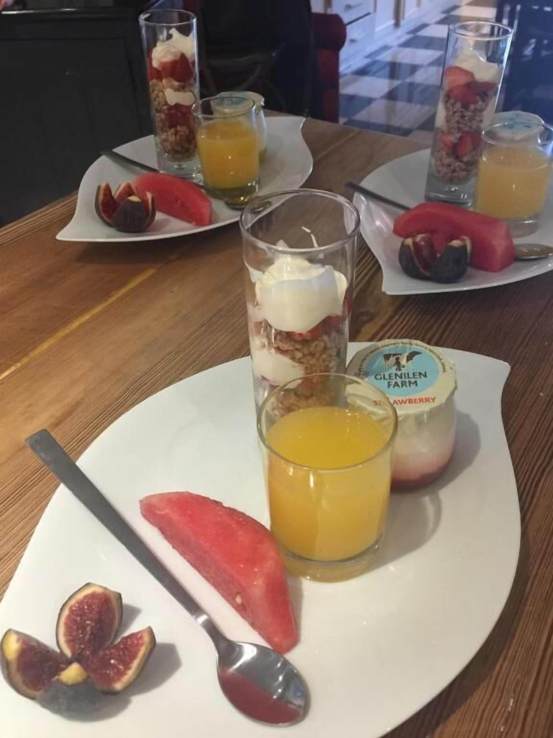 Une table mise pour le petit-déjeuner avec une assiette de fruits, un verre de jus d’orange et un pot de yaourt.