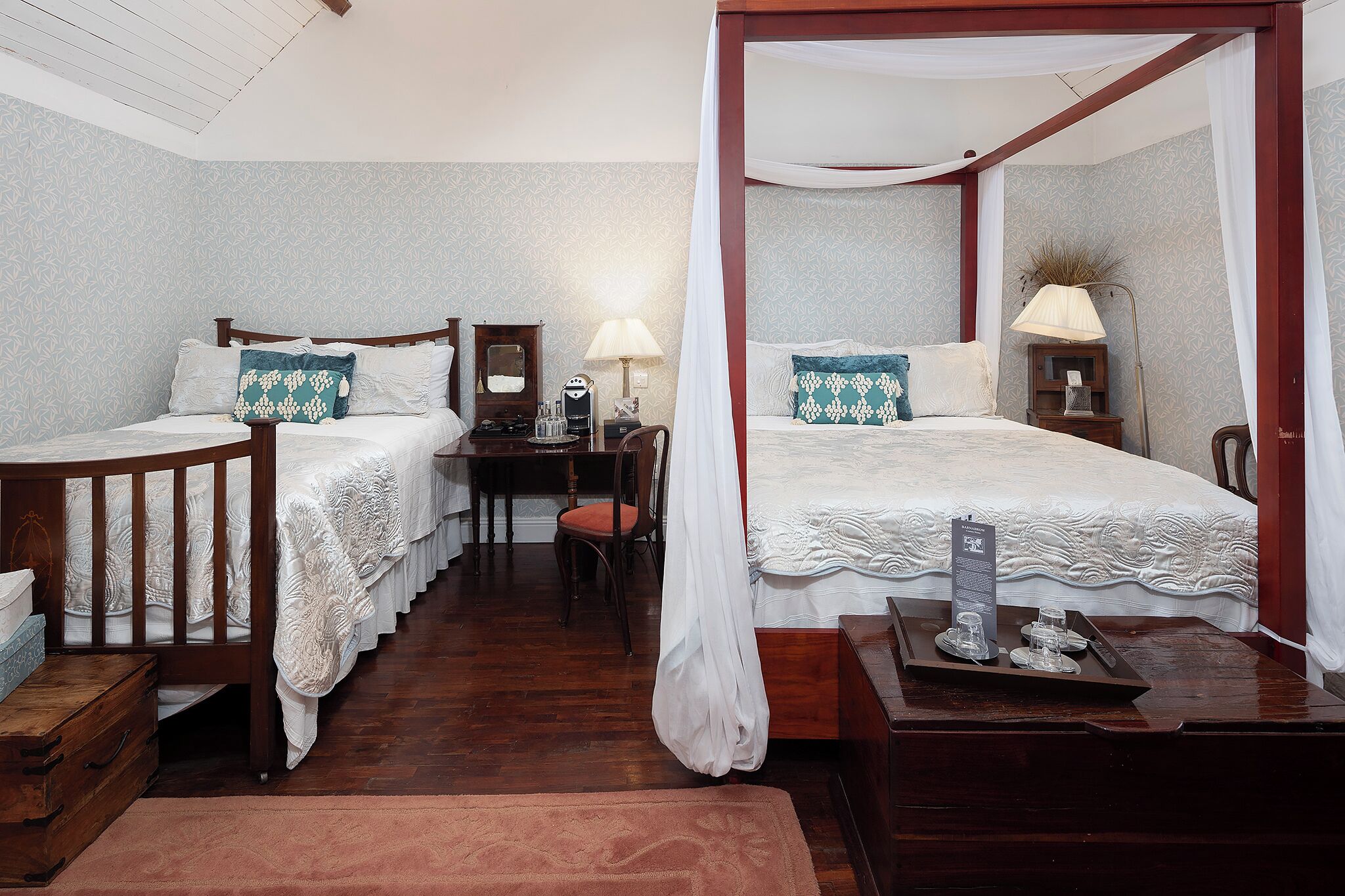 A four-poster bed with a canopy, a desk with a chair, a nightstand with a lamp, and a wooden chest of drawers.