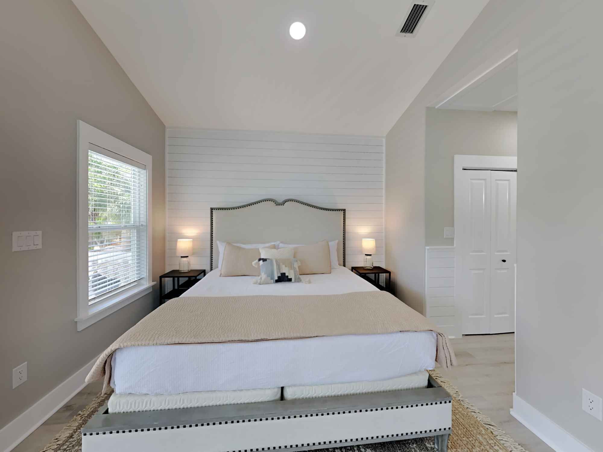 A bedroom with a large bed, two bedside tables with lamps, a window with blinds, and a door.