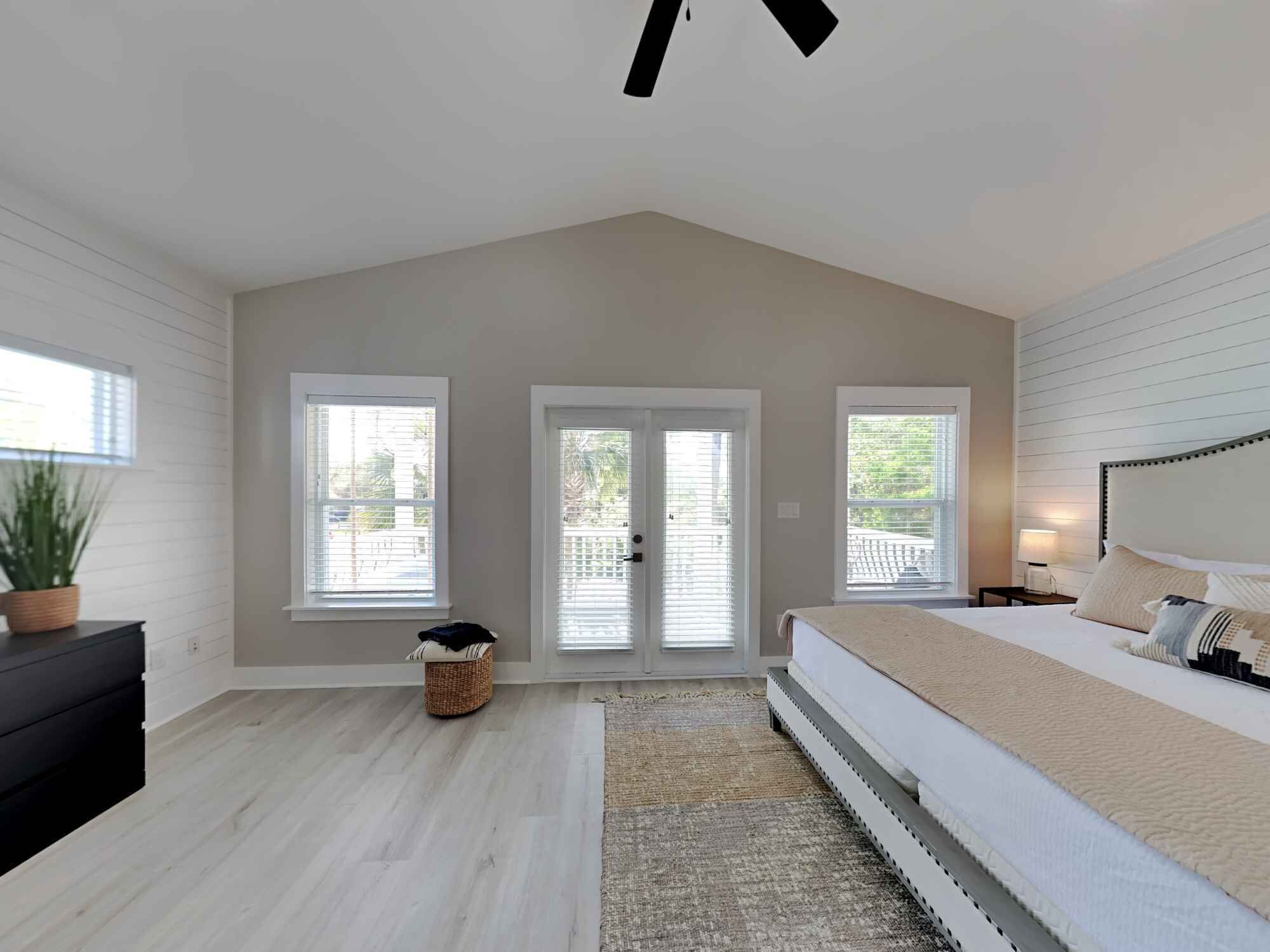 A bedroom with a large bed, two windows, a ceiling fan, and a small side table.