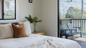 A bedroom with a bed, pillows, a bedside table with a plant, and a balcony with chairs.