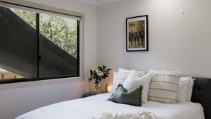 A bedroom with a large window, a framed picture of The Beatles on the wall, a bedside table with a vase of flowers, and a bed with white bedding and green pillows.