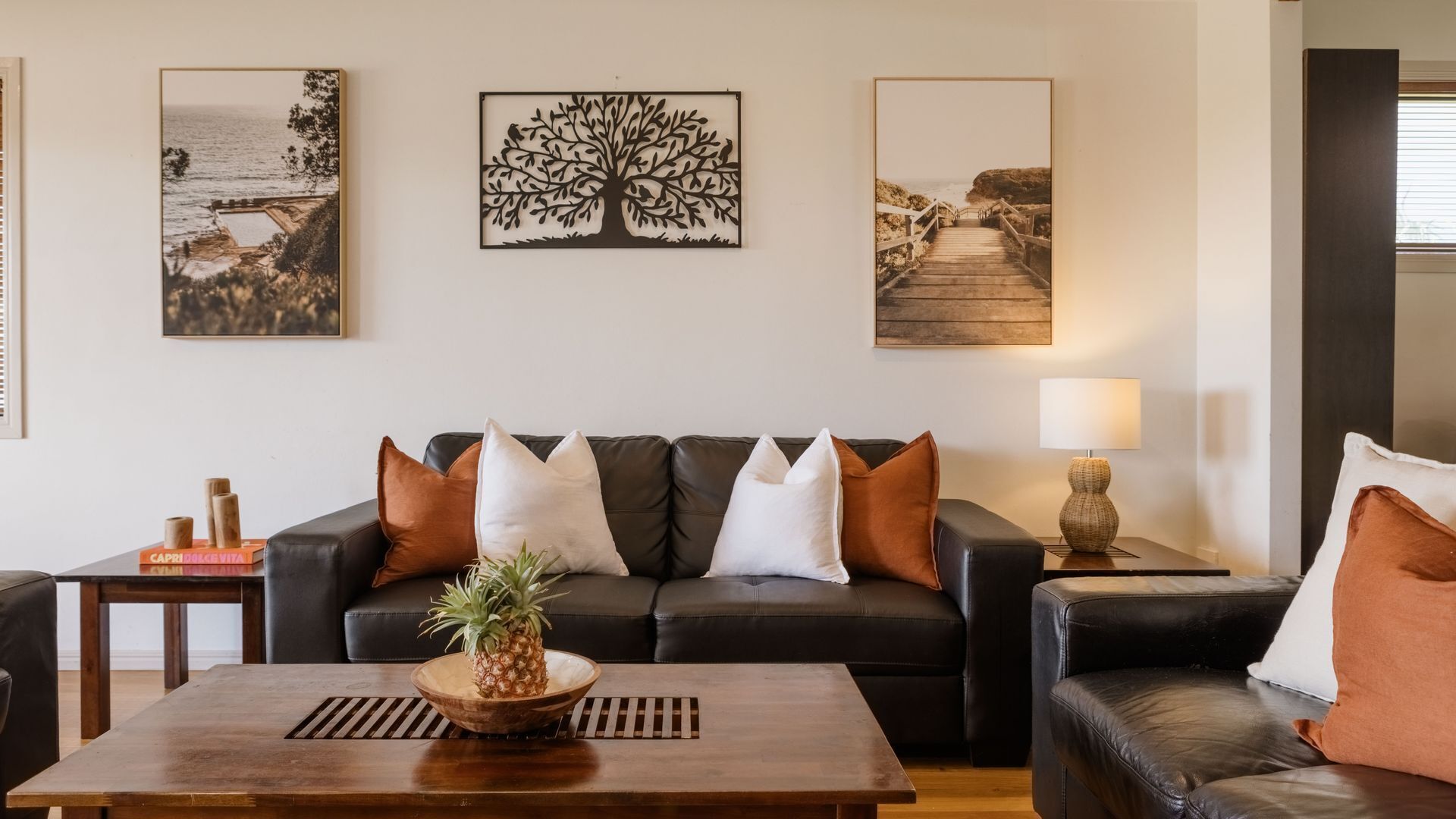 A living room with a dark leather sofa, a wooden coffee table with a pineapple centerpiece, and three framed artworks on the wall.