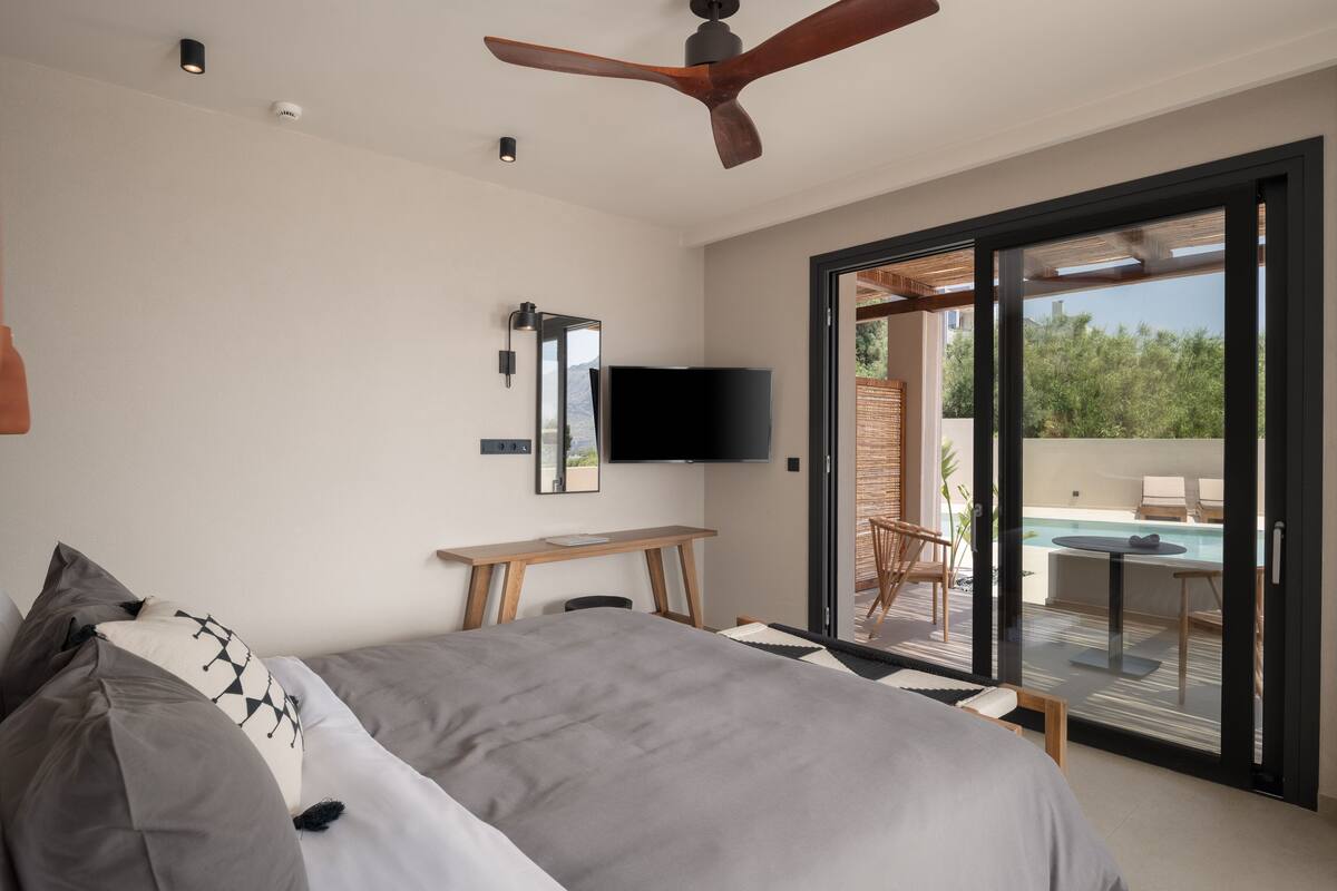 Un dormitorio con cama, escritorio, televisor y puerta corrediza de vidrio a un área exterior con piscina.