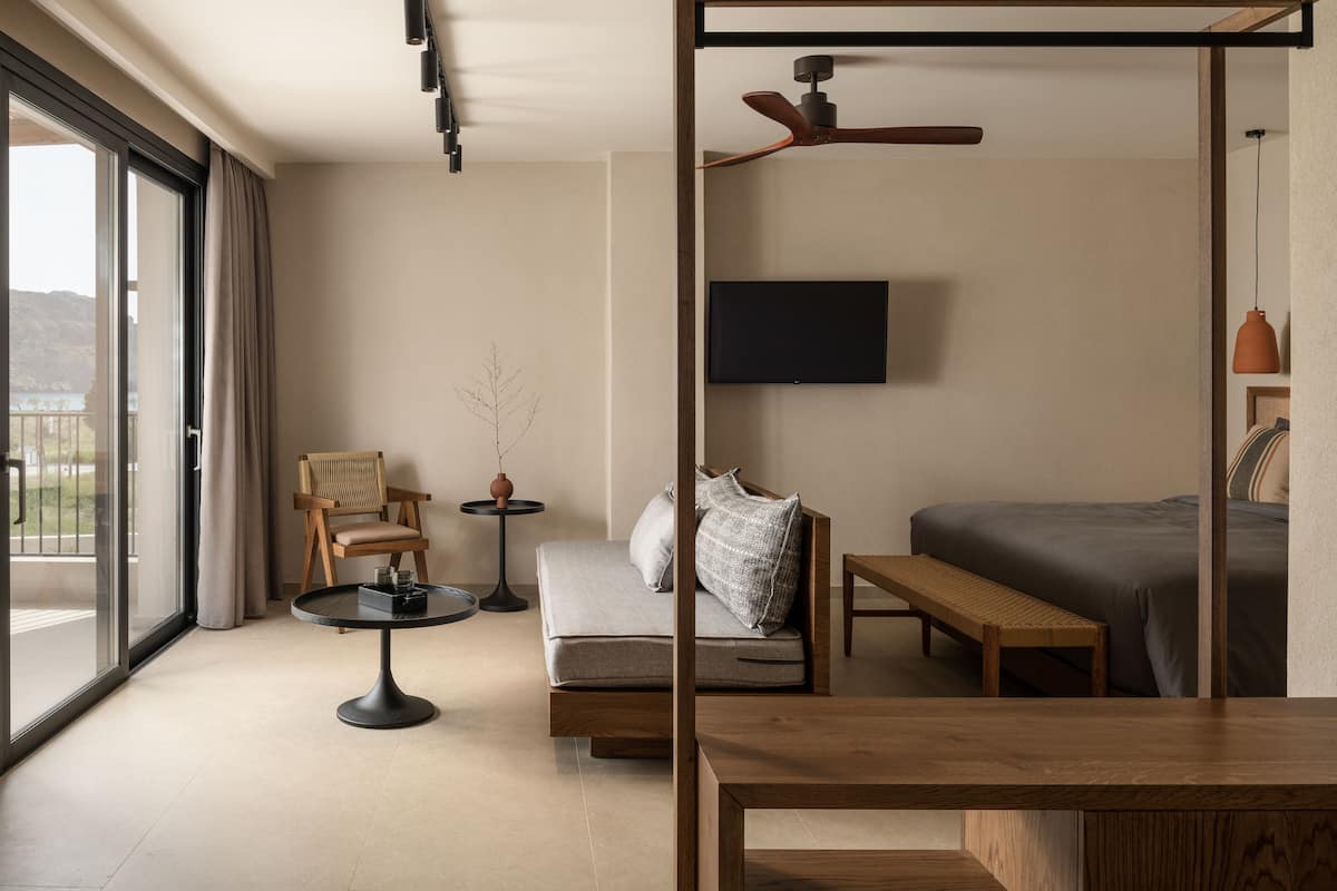 Habitación de hotel moderna con un ventanal grande, una cama, un sofá, una mesita, un sillón, un televisor y un ventilador de techo.